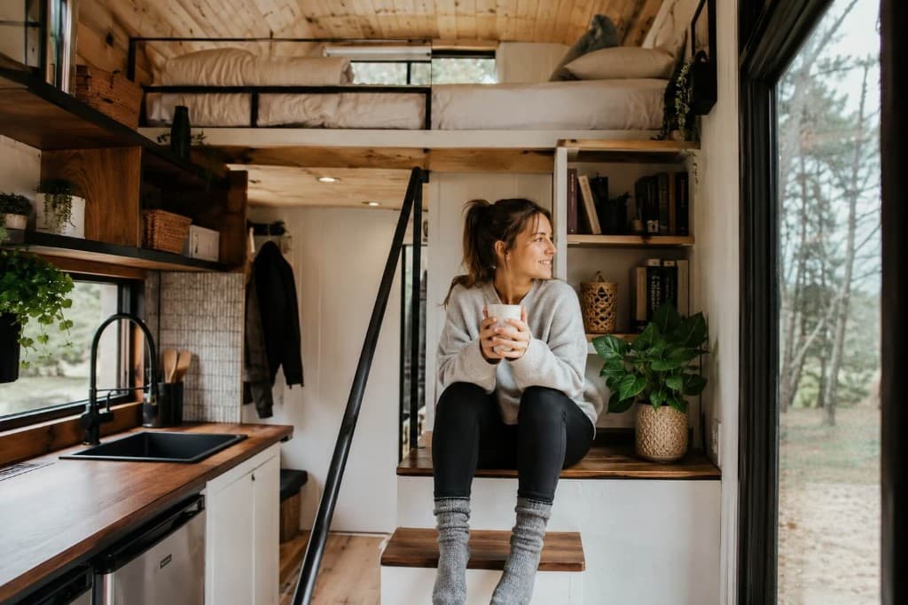 Woman relaxing with coffee inside a cozy modern tiny house with kitchen, loft bed, and natural light